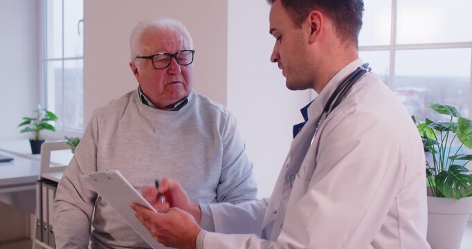 Male young doctor examining senior elderly man sitting on couch in exam room during medical checkup in clinic. Man physician talking with mature patient in office. Medicine concept. 4k video.