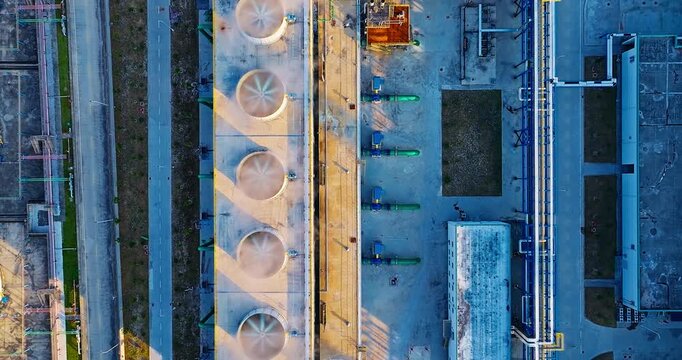 Top-down aerial view of industrial cooling towers with rotating fans, pipelines, and factory infrastructure during sunset.