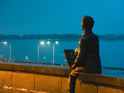 Volgograd, Russia &ndash; July 5, 2023: Sculpture. A man reads a book while sitting on the embankment parapet at dusk, set against the backdrop of city lights