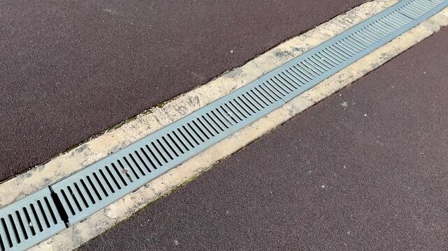 Galvanized drainage grate along stadium curb, long metal channel between asphalt and concrete, repeating slot pattern, leading