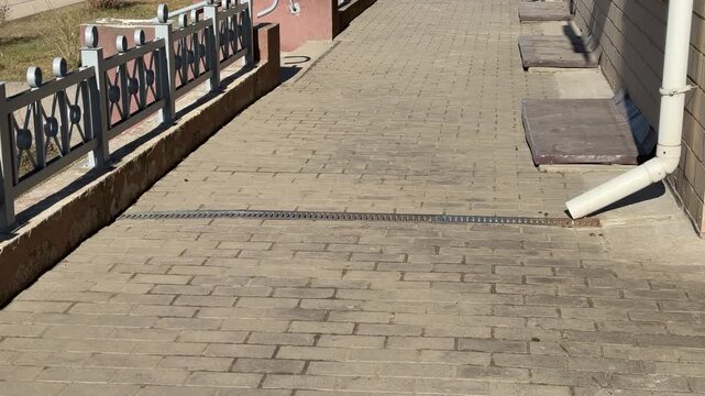 White pipe metal grate on pavement, patterned metal grate covers spillway installed across paving stones on footpath, sunlight