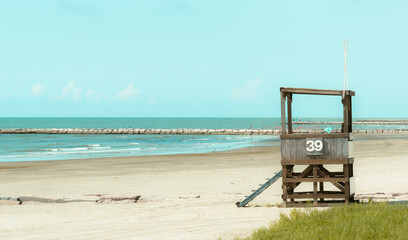 GALVESTON, TEXAS, USA: A wooden beach lifeguard tower displaying the number 39 stands on an empty sandy beach with a stone breakwater jetty and the Gulf of Mexico in the background © YuliiaK