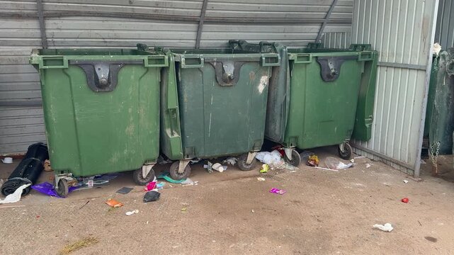 Green dumpsters with scattered trash under bright summer sunlight, gravel foreground, worn metal bins, colorful packaging