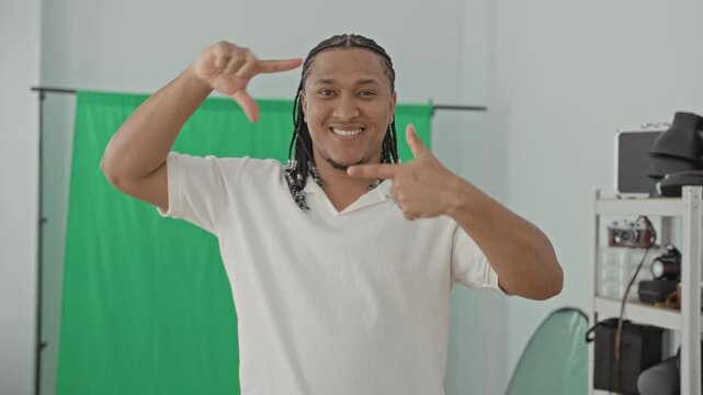 Man frames a camera shot with hands and smiles in front of a green screen backdrop in a studio filled with camera gear and reflectors; playful creativity.