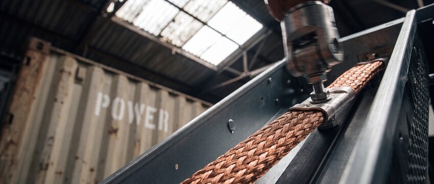 Braided copper grounding cable connection secured to industrial steel frame in power facility warehouse, representing high voltage electrical safety, heavy industry infrastructure, and energy systems.