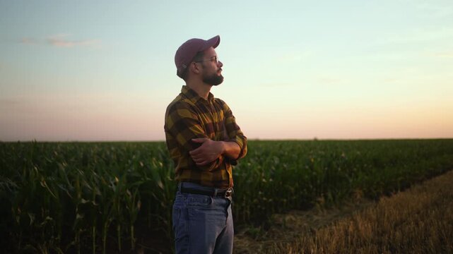 A farmer watches the sun setting in a wide field. He wears a hat and flannel shirt. Crops grow on one side of the field, while the other side is harvested.