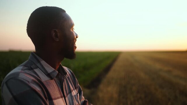 An Afro American Farmer stands in a field with a smile on his face as the sun sets in the background. The sky shows warm colors. 