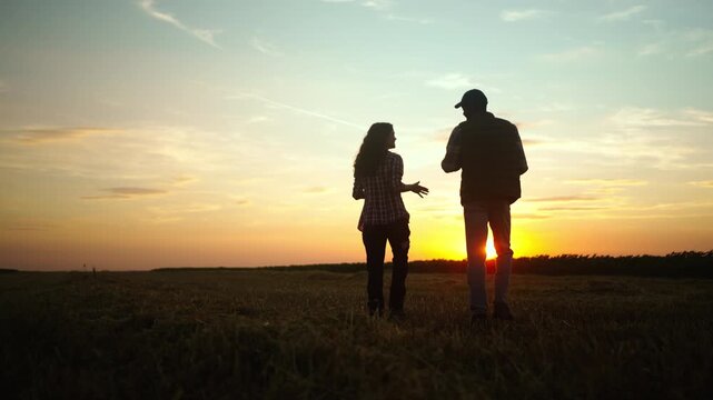 Two farmers walk side by side in a field during sunset. They enjoy the view as the sun lowers on the horizon. The sky shows colors of orange and yellow.