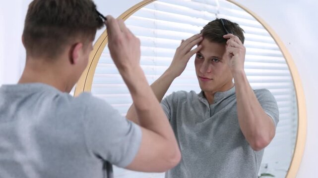 Handsome young man brushing his hair with comb near mirror in bathroom