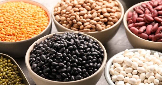 Different types of legumes in bowls on grey table, closeup. Camera moving right