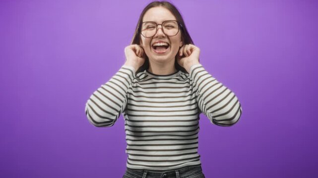 Woman wearing glasses and striped sweater touching her ears and smiling in studio with purple backdrop; joy.