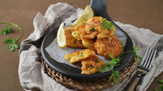 Crispy cod fish fritters are arranged in a rustic black skillet on a woven mat, accompanied by lemon wedges and fresh parsley. Warm studio lighting highlights the textures and golden-brown crust of th