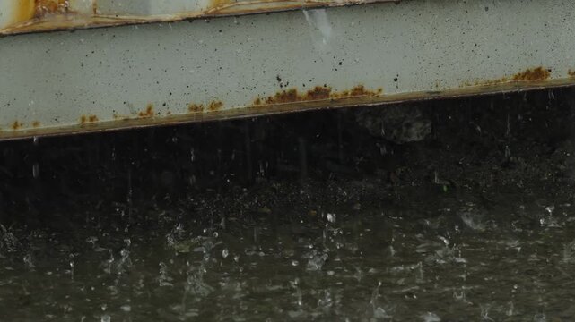 Rainwater dripping from a rusty metal edge into large puddles.