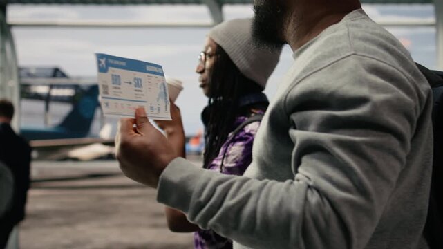 Close up of airplane ticket used by people walking in airport terminal traveling together to foreign countries, African American man and woman reading boarding pass flight information.
