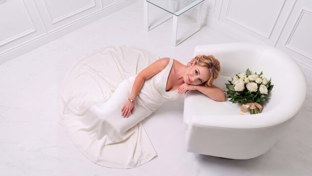 A bride in a white satin gown lounges on a white marble floor, resting against a modern armchair beside a white rose bouquet. Minimalist composition.