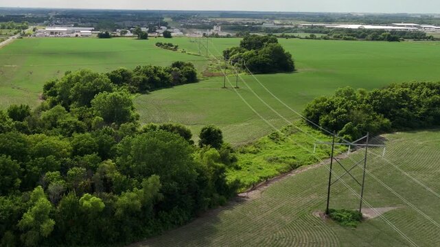 Aerial view of electrical utility transmission lines crossing rural farmland cornfields toward a distant industrial zone and technology park
