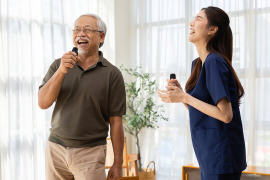 Happy Asian senior man and friendly nurse singing karaoke with microphone device inside bright living room, laughing together during joyful recreation therapy session with caregiver