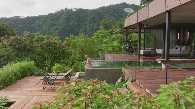 Girl swims in infinity pool at luxurious villa surrounded by lush tropical rainforest mountains. Modern architecture with wooden deck and loungers creates serene resort atmosphere for relaxation.