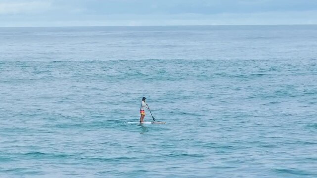 A lone paddleboarder glides across vast turquoise ocean waters under an overcast sky, wearing colorful shorts and a cap while navigating gentle rippling waves on a calm peaceful day at sea.