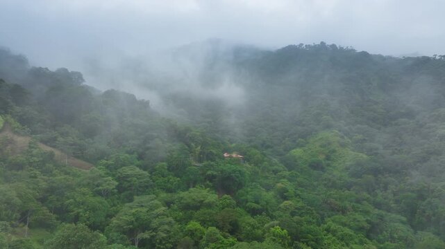 Aerial view of Costa Rica rainforest covered mountains with thick mist and fog rolling through dense tropical vegetation. Small houses with red roofs nestled among the lush green jungle canopy.