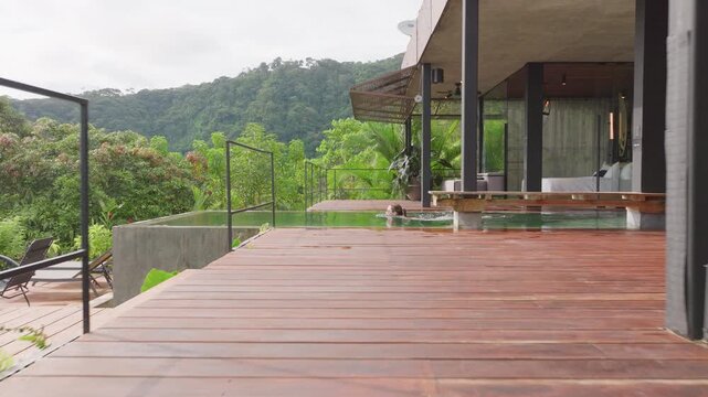 Woman swimming in infinity pool at modern luxury villa with wooden deck, concrete architecture, and glass walls overlooking lush green tropical rainforest mountains under overcast skies.