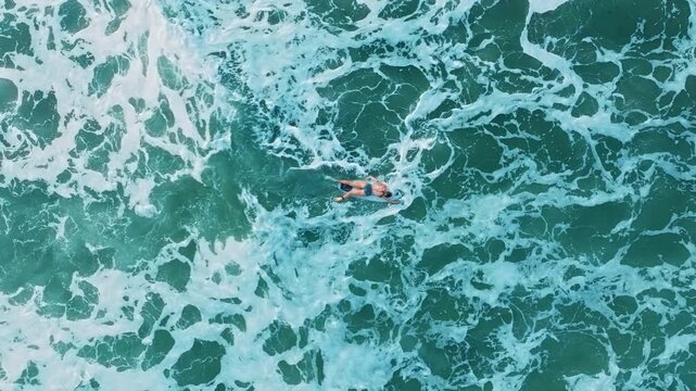 Surfing adventure captured from above as swimmer navigates through stunning turquoise ocean waters. White foam patterns create mesmerizing texture around the person floating in tropical sea waves.