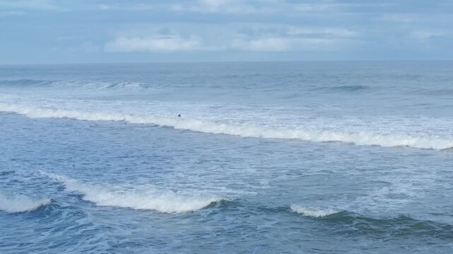 Surfing scene captures lone surfer paddling through rolling ocean waves under cloudy skies. White foam crests as multiple wave lines break across blue water with rocky coastline visible in distance.