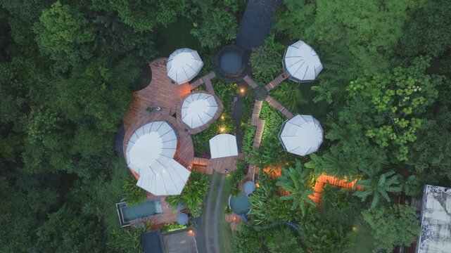 Beautiful villa bungalow in Costa Rica features white geodesic dome structures connected by wooden walkways amid dense tropical rainforest. Aerial view reveals pool and evening lights.