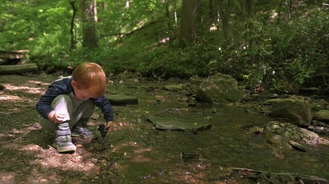 Young boy with light brown hair crouches down beside a stream in a forest, extending his hand towards a toy car resting in the water, with rocks and lush greenery surrounding him