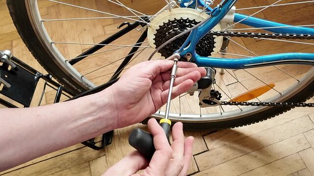Close-up of hands adjusting bicycle derailleur with a screwdriver on a wooden floor. A person is meticulously working on a bicycle's rear derailleur system, using a screwdriver to make adjustments to 