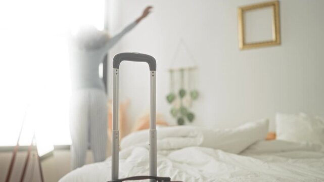 Woman stretches arms toward a bright window with suitcase handle prominent in foreground inside building, beside an unmade bed and macrame wall hanging; anticipation travel.