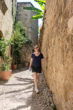 Mujer joven y sonriente caminando por un estrecho callej&oacute;n empedrado de una ciudad hist&oacute;rica. Muros altos de piedra, sombras proyectadas y plantas adornan el pintoresco paseo.