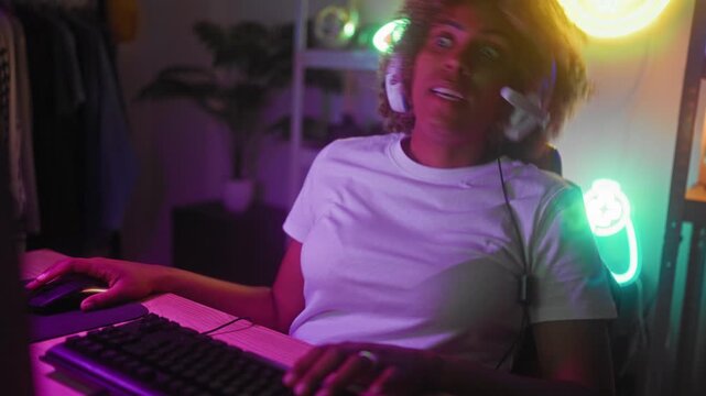 Young african american woman wearing headset, holding microphone and hand on mouse, smiling while playing on computer in studio; joy connection.