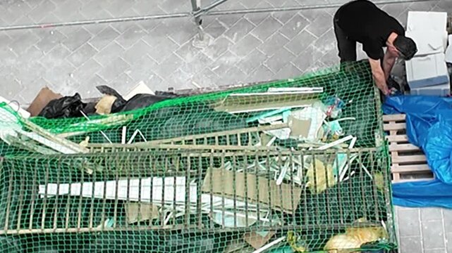 Construction site worker managing waste materials in a dumpster with safety gear. A worker in protective gloves sorts debris in a construction site dumpster, highlighting waste management and safety