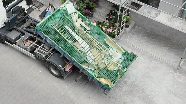 Construction debris removal truck with scaffolding and plants on pavement. A truck is parked on a paved surface, its dumpster filled with construction debris and covered by a green net, with scaffoldi