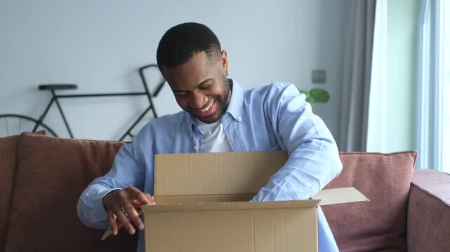 Young happy man sitting on the couch unboxing a cardboard box with a satisfied and curious expression