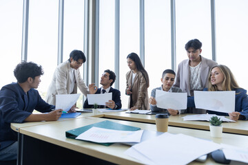 Fototapeta premium Diverse group of young professionals sitting at large conference table holding documents feeling focused and engaged during important modern corporate team meeting in bright glass walled office