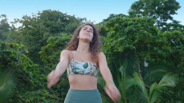 Athletic woman with curly hair performs warm up before surfing exercises outdoors. She stretches arms and moves gracefully in floral crop top against lush green tropical foliage and palm trees.