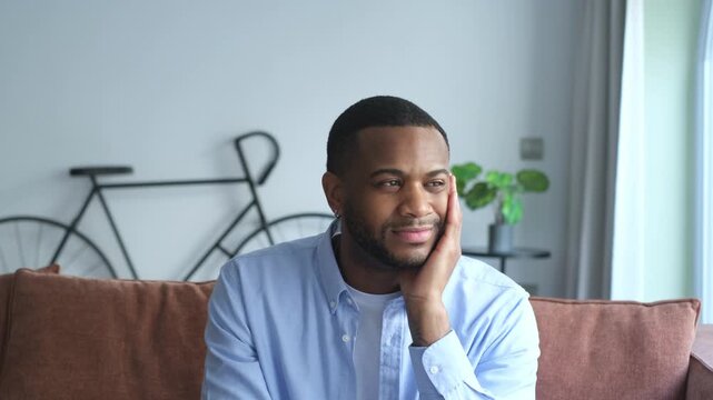 Handsome young man sitting on couch, looking thoughtfully out the window before smiling at camera
