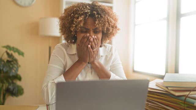 Woman at laptop with hands covering mouth and fingers clasped, seated at desk with books and plant in a building; anxiety work pressure.