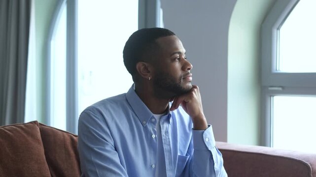 Pensive young Black man sitting on a sofa and looking thoughtfully out the window at home