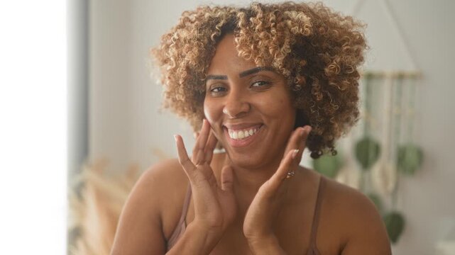 Woman smiling and touching face with hands framing cheeks, visible bra straps, indoor portrait set with macrame decor in studio; self care joy.