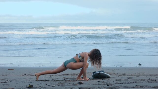 Young woman in bikini performs warm-up stretches before surfing on sandy beach. She does lunges and forward bends next to her surfboard while ocean waves roll in the background under cloudy sky.
