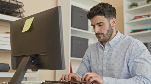 Man massaging his wrist while seated at a desktop computer and typing on a keyboard, sticky note on monitor and shelves with storage boxes visible in building; strain focus productivity.