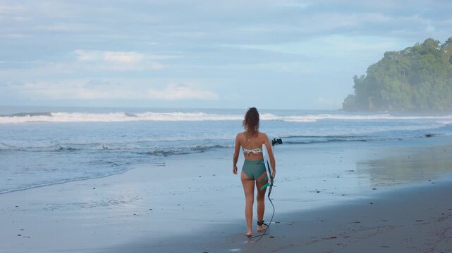 Young woman in green bikini walks barefoot on dark sand beach carrying white surfboard toward breaking waves. Tropical forested headland rises in misty background under cloudy sky.