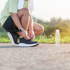 Obraz premium Asian man with towel tying running shoes in the park with a reusable water bottle. male runner getting ready for jogging and fitness exercise. Healthy lifestyle and sustainability concept