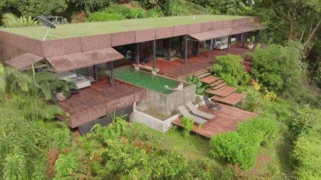 Aerial drone footage of modern architectural villa with green roof and wooden deck terraces nestled in lush tropical jungle as woman relaxes swimming in concrete infinity pool with waterfall feature.