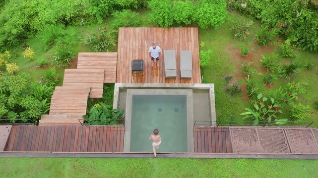 Aerial drone view of a young boy jumping into a modern concrete plunge pool while his father watches from a wooden deck with lounge chairs, surrounded by lush tropical greenery and gardens.