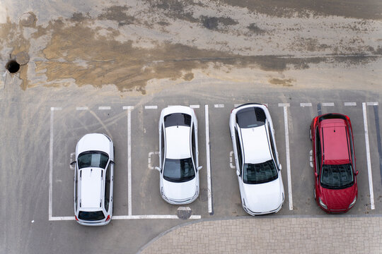 Top view of white and red cars parked in designated parking lots on asphalt near a dirty road surface with an open manhole hatch.