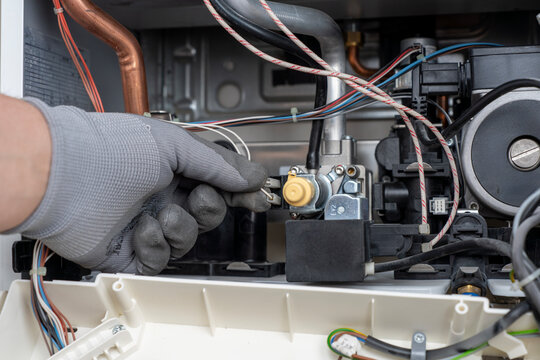 Close-up of a technician hand in a protective glove connecting a wire terminal to a gas valve modulator inside an open domestic boiler during maintenance.